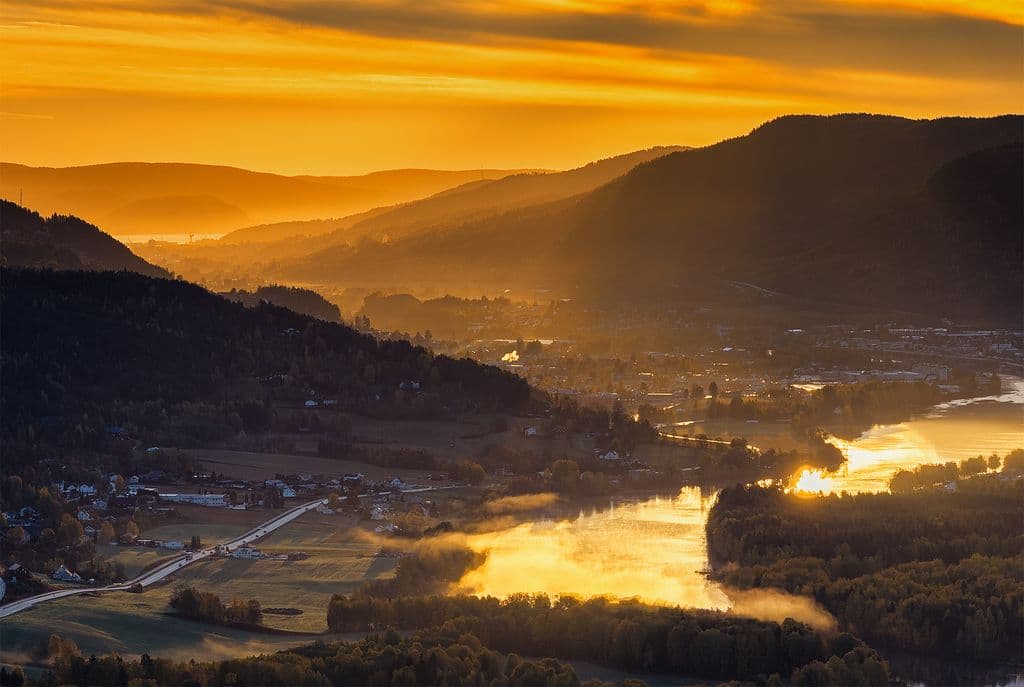 A long-lens aerial capture from above Hokksund, looking east through the Drammenselva valley toward Mjøndalen and the port of Drammen.